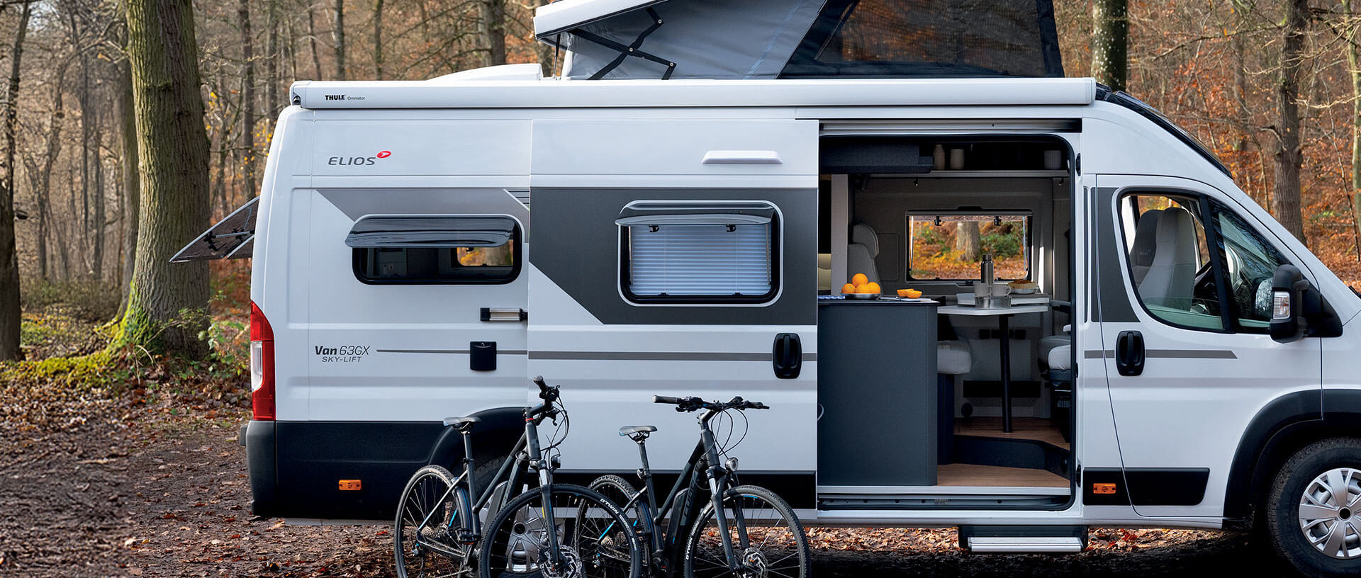 Camper Elios 63GX Sky Lift con techo elevable, puerta lateral abierta y bicicletas en el exterior